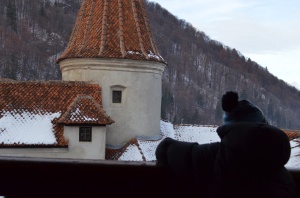 A child in silhouette looks out at Bran Castle, white with a red roof and red turret, and a snowy, treed mountain behind it.